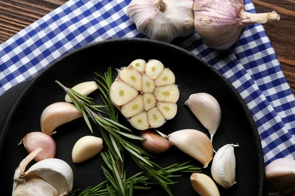 Fresh garlic with rosemary in black pan near blue checkered cotton napkin on wooden background, close up