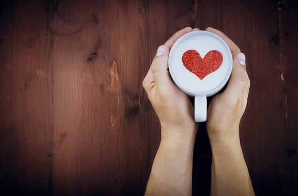 Man holding hot cup of milk on wood table, with red heart shape
