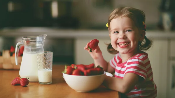 Happy child girl eats strawberries in summer home kitchen