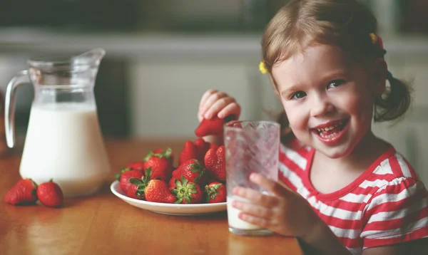 Happy child girl drinks milk and eats strawberries in summer hom