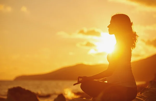 Pregnant woman practicing yoga in lotus position on beach at sun
