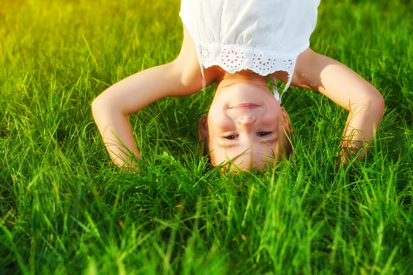 Happy child girl standing upside down on his head on grass in su