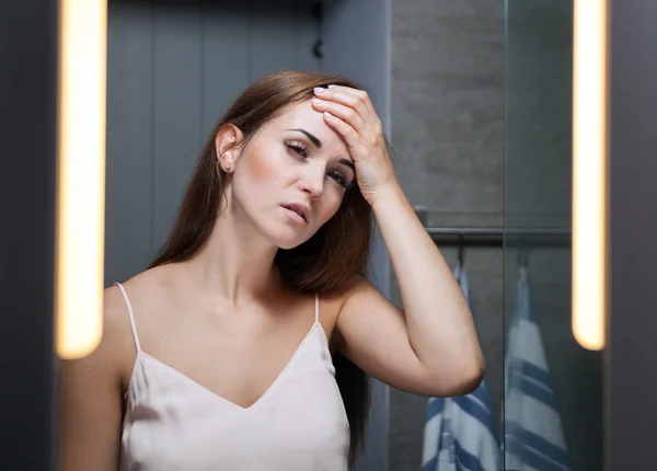 Woman with headache in front of a bathroom mirror