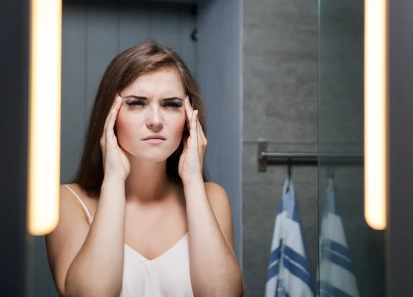 Woman with headache in front of a bathroom mirror