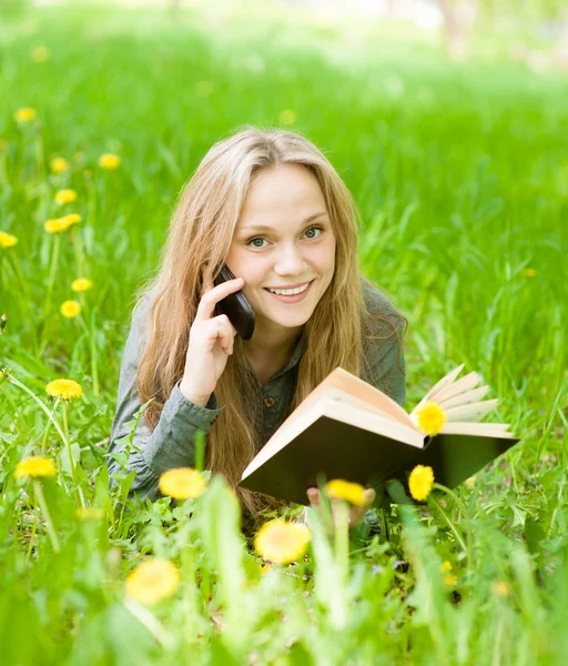 Girl lying on grass with dandelions reading a book and talking on phone