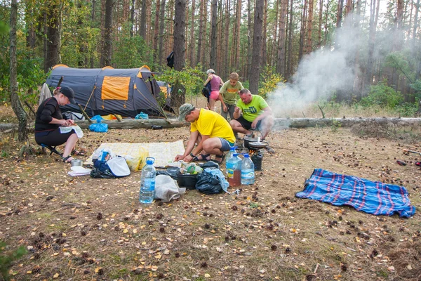 A group of tourists set up a tent in the woods and cook a meal on a campfire