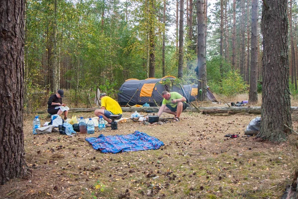 A group of tourists set up a tent in the woods and cook a meal on a campfire