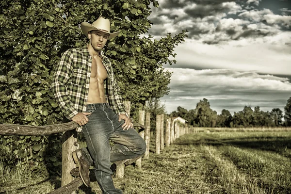 Portrait of rustic man in hat with unbuttoned shirt leaning on wooden fence