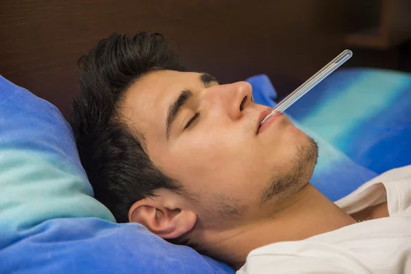 Young man in bed measuring fever with thermometer
