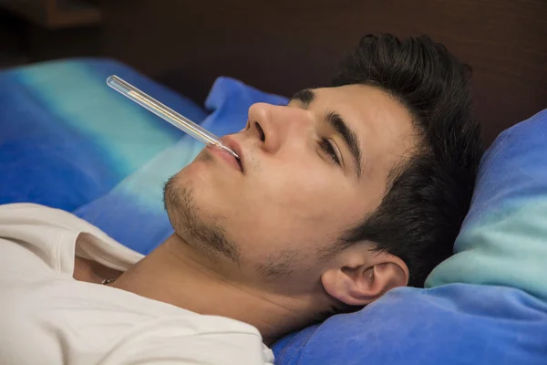 Young man in bed measuring fever with thermometer