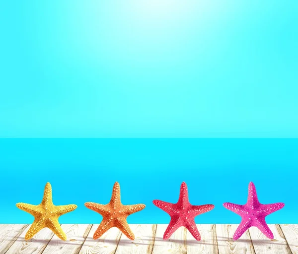Yellow, orange, red and pink sea stars standing in a row on white wooden surface of the pier or Wharf. Behind there is a sea background. Blue cloudless sky and warm blue sea.