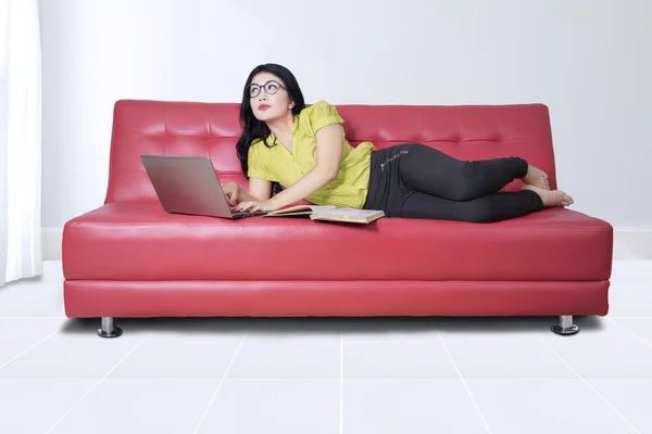 Woman with book and notebook computer on sofa