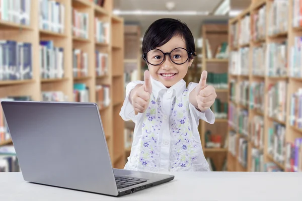 Child with laptop giving thumbs up in library