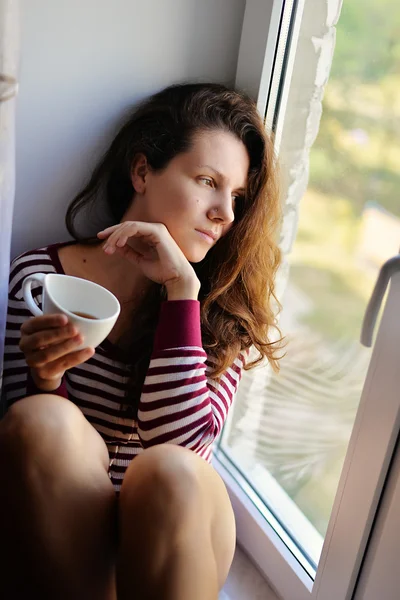 The sad girl sits at a window with a cup of tea, coffee