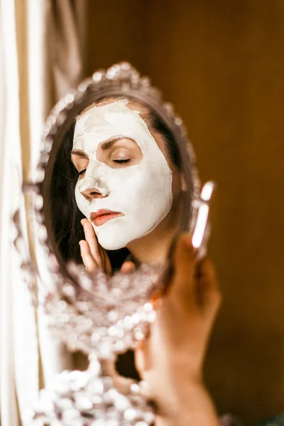 Woman applying mask of clay skin on face looking in mirror.