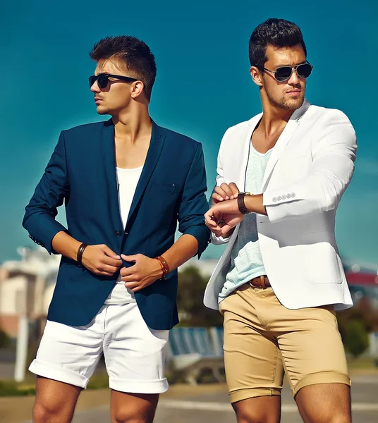 Fashion portrait of two young sexy handsome men models in casual cloth suit in sunglasses in the street behind blue sky