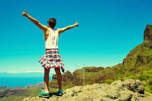 Happy stylish man in casual hipster clothes standing on the cliff of mountain with raised hands to the sun and celebrating success