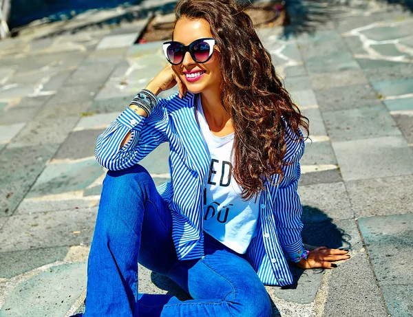 Fashion stylish beautiful young smiling brunette woman model in summer hipster casual blue clothes sitting in the street in sunglasses