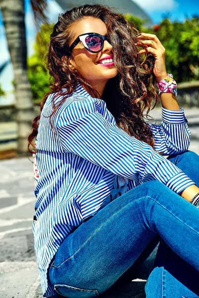 Fashion stylish beautiful young smiling brunette woman model in summer hipster casual blue clothes sitting in the street in sunglasses