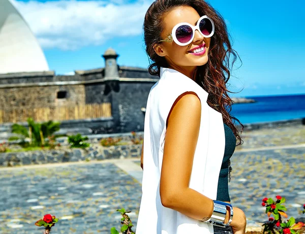 Portrait of beautiful smiling sexy brunette businesswoman model in white clothes in sunglasses posing behind blue sky