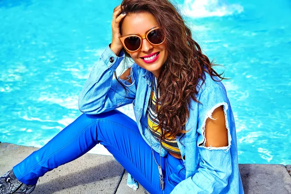 Fashion stylish beautiful young smiling brunette woman model in summer  hipster clothes posing in the street behind blue water in sunglasses
