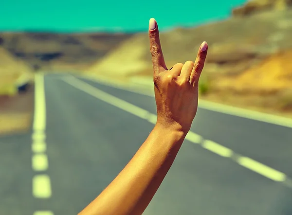 Young caucasian tourist hitchhiking along a road in sunset and showing rock and roll sign