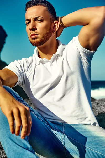 Young stylish handsome man model in hipster summer clothes posing on beach behind sky and ocean