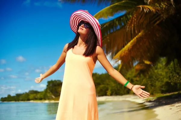 Hot beautiful woman in colorful sunhat and dress walking near beach ocean on hot summer day near palm