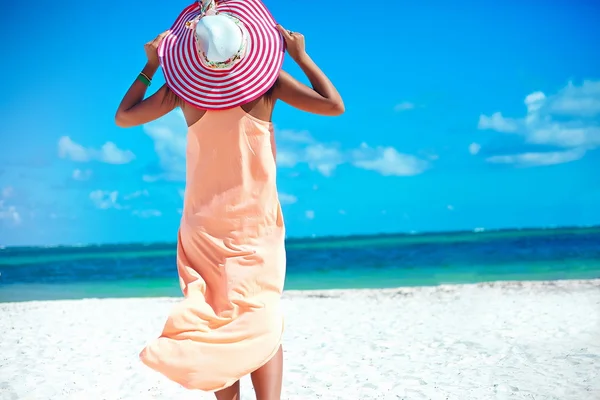 Hot beautiful woman in colorful sunhat and dress walking near beach ocean on hot summer day on white sand