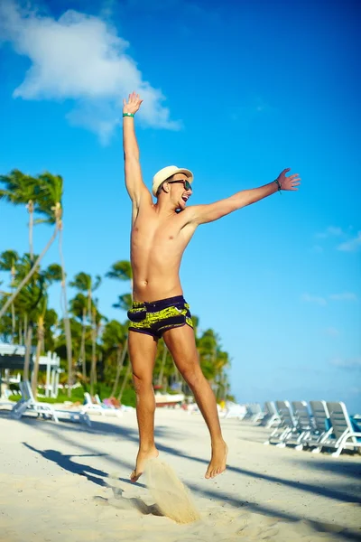 Happy handsome muscled man in sunhat on beach jumping behind blue sky behind blue sky