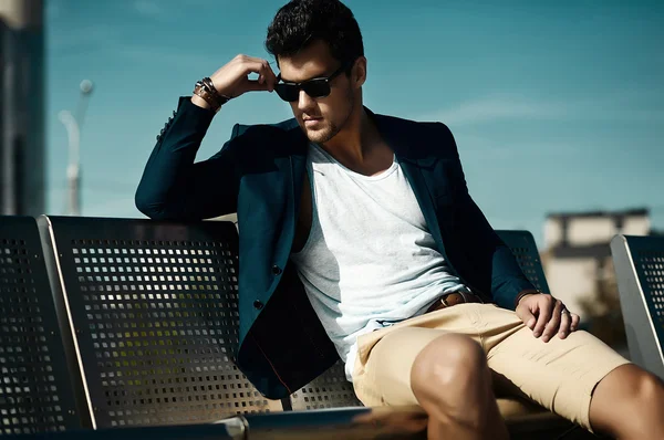 Fashion portrait of young sexy businessman handsome model man in casual cloth suit in sunglasses sitting on a bench in the street