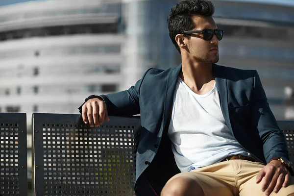 Fashion portrait of young sexy businessman handsome model man in casual cloth suit in sunglasses sitting on a bench in the street