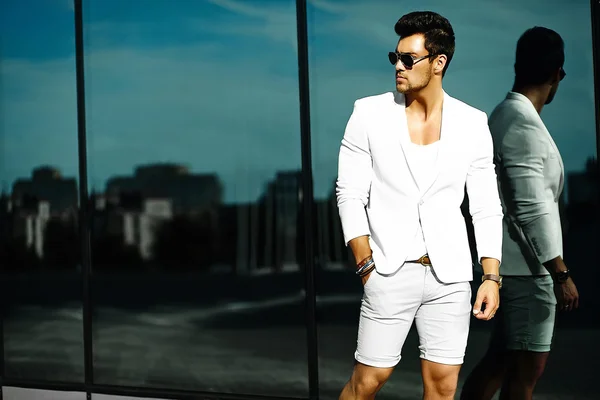 Fashion portrait of young sexy handsome model man in casual white suit in sunglasses in white suit in the street