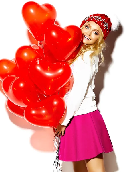 Portrait of beautiful happy sweet cute smiling blonde woman girl in casual hipster clothes, in pink skirt and winter warm beanie with red heart balloons in hands