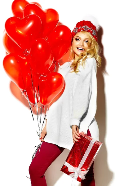 Portrait of beautiful happy sweet smiling blonde woman girl holding in her hands big Christmas gift box and heart balloons in casual red hipster winter clothes, in white warm sweater