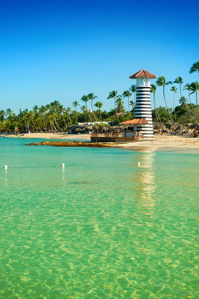 Striped lighthouse on sandy shore with palm trees. Clear water of the Caribbean sea.