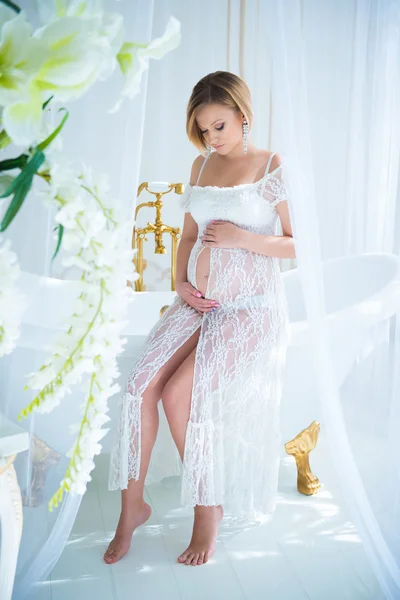 Pregnant girl in a white lace dress sitting in the interior with flowers and hugs belly