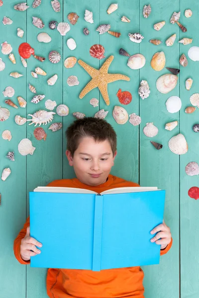 Cute boy reading blank book near seashells