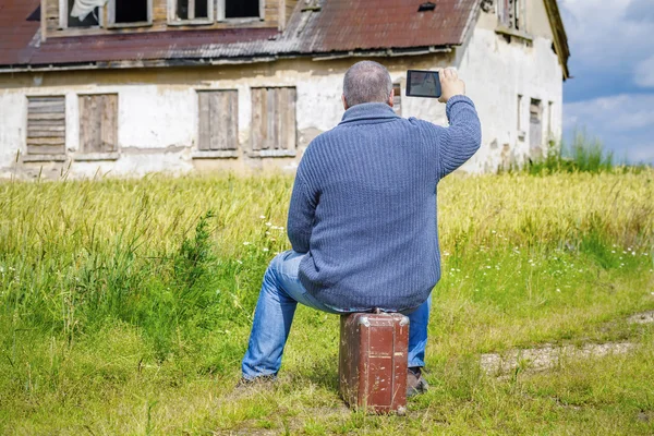 Man sitting on suitcase and take pictures of old house