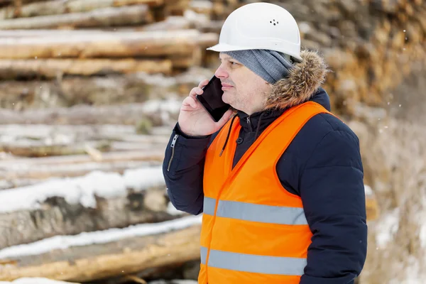 Worker with smartphone near piles of logs in winter