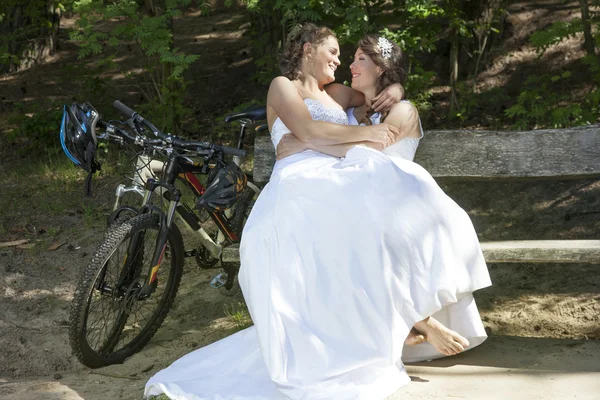 Two brides on bench in forest with mountain bikes