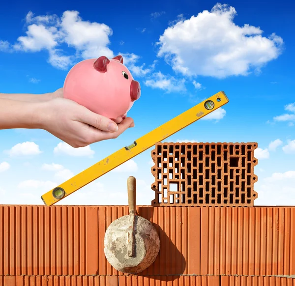 Masonry tools on a brick wall and a hand holding a piggy bank.