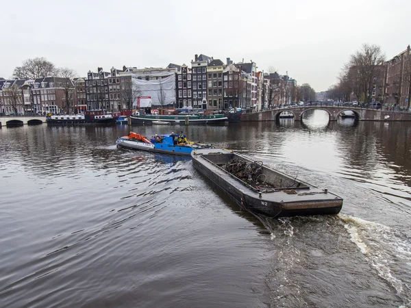AMSTERDAM, NETHERLANDS on MARCH 31, 2016. Typical urban view. A row of traditional architecture on the canal embankment. The barge loaded with the sunk bicycles floats on the channel. Houseboats near the banks