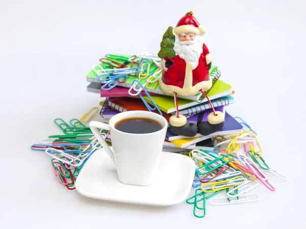 Rough pile of notebooks in covers of various colors, multi-colored paper clips, a cup of black coffee and Father Frost\'s figure