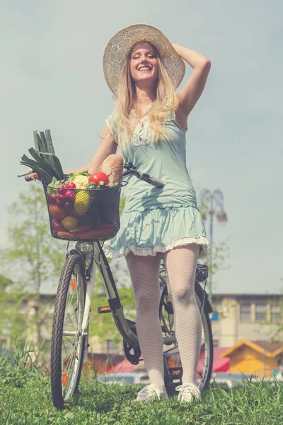 Attractive blonde woman with straw hat posing next to bike with basket  full of groceries.