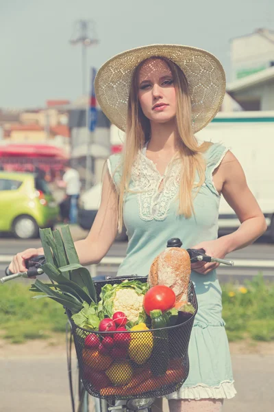 Attractive blonde woman with straw hat posing next to bike with basket  full of groceries.