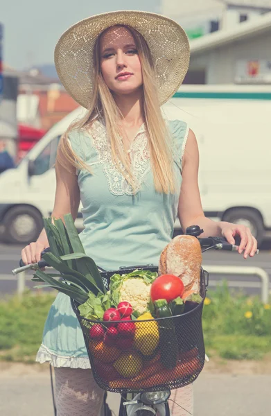 Attractive blonde woman with straw hat posing next to bike with basket  full of groceries.