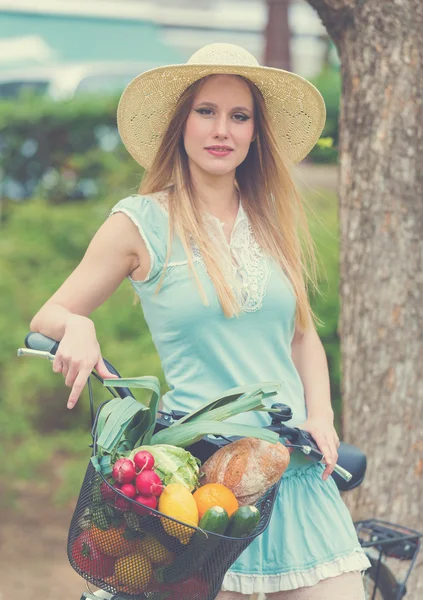 Attractive blonde woman with straw hat standing in the park and posing next to bike with basket full of groceries.