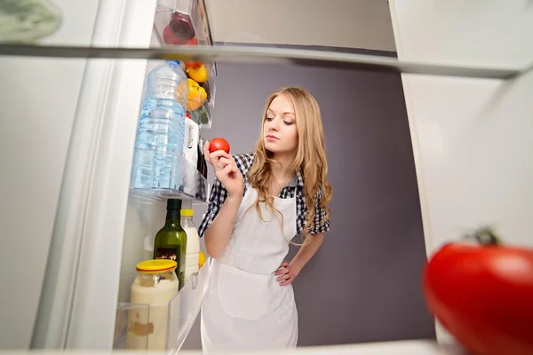 Blonde woman pulls tomato from the refrigerator.