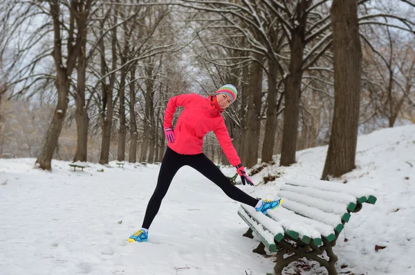 Winter running in park: happy woman runner warming up and exercising before jogging in snow, outdoor sport and fitness concept